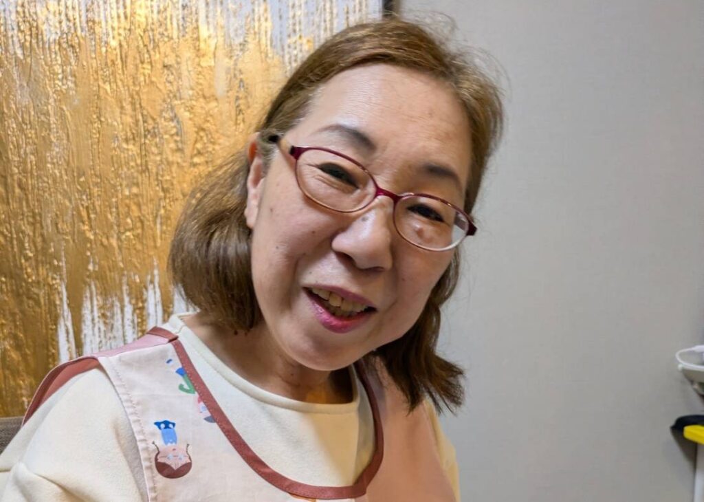 Smiling elderly woman with short brown hair and red glasses wearing a beige apron, in a room with a gold textured wall behind her.